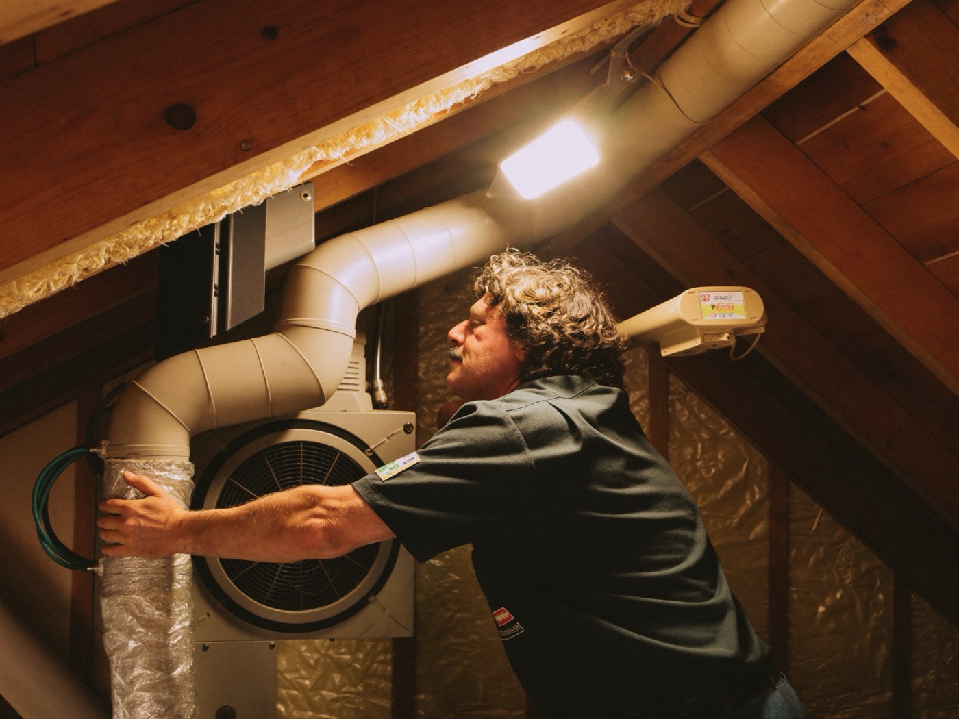 Technician in a green work shirt servicing a furnace in a cramped attic by work light